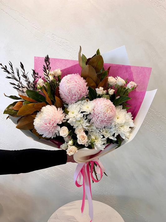 A beautiful foward facing bouquet with pink mums, spray roses and white daisies with lush foliage presented in a wrap and finished with ribbons.
