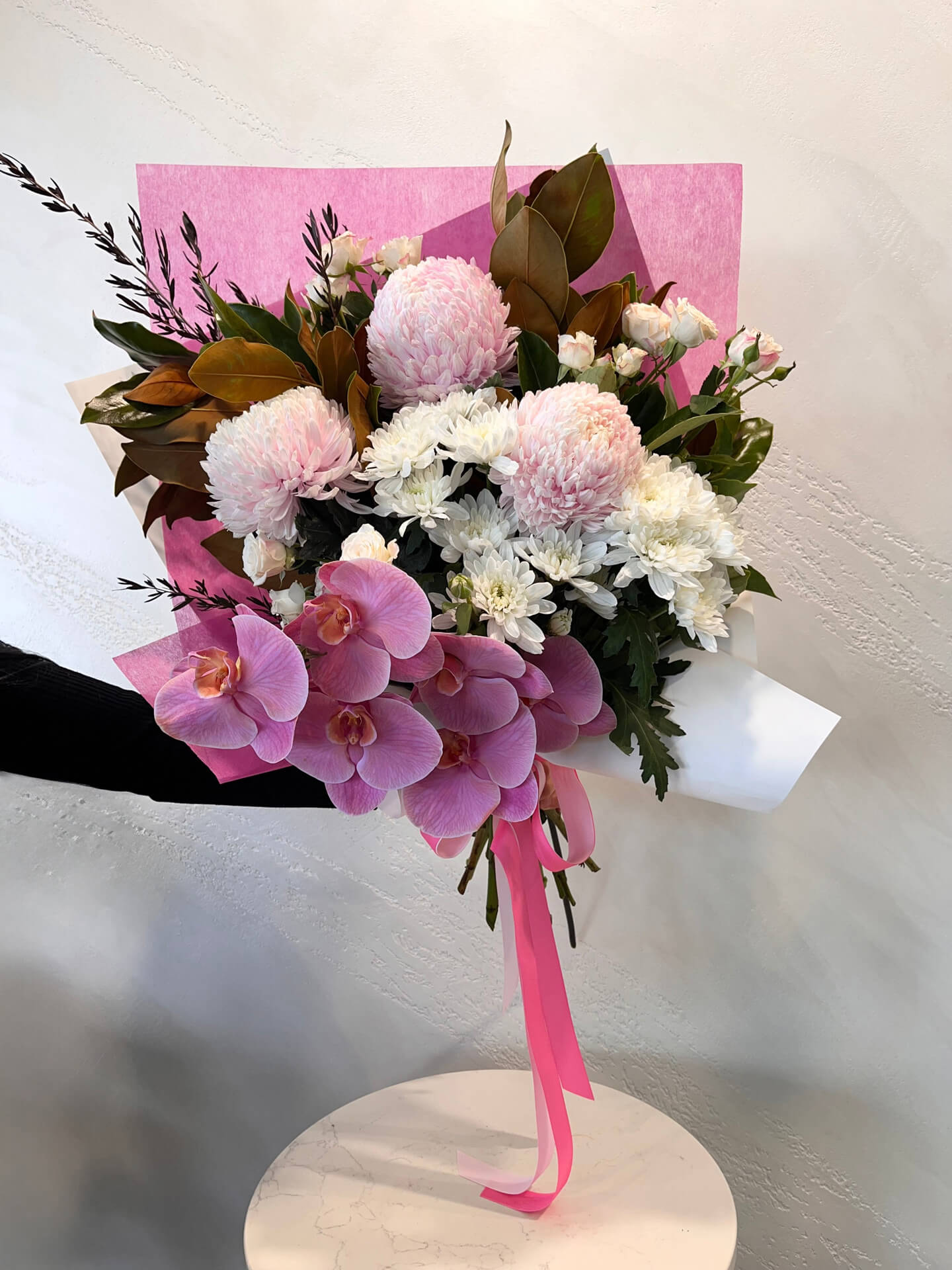 A beautiful foward facing bouquet featuring a phalaenopsis orchid with pink mums, spray roses and white daisies with lush foliage presented in a wrap and finished with ribbons.