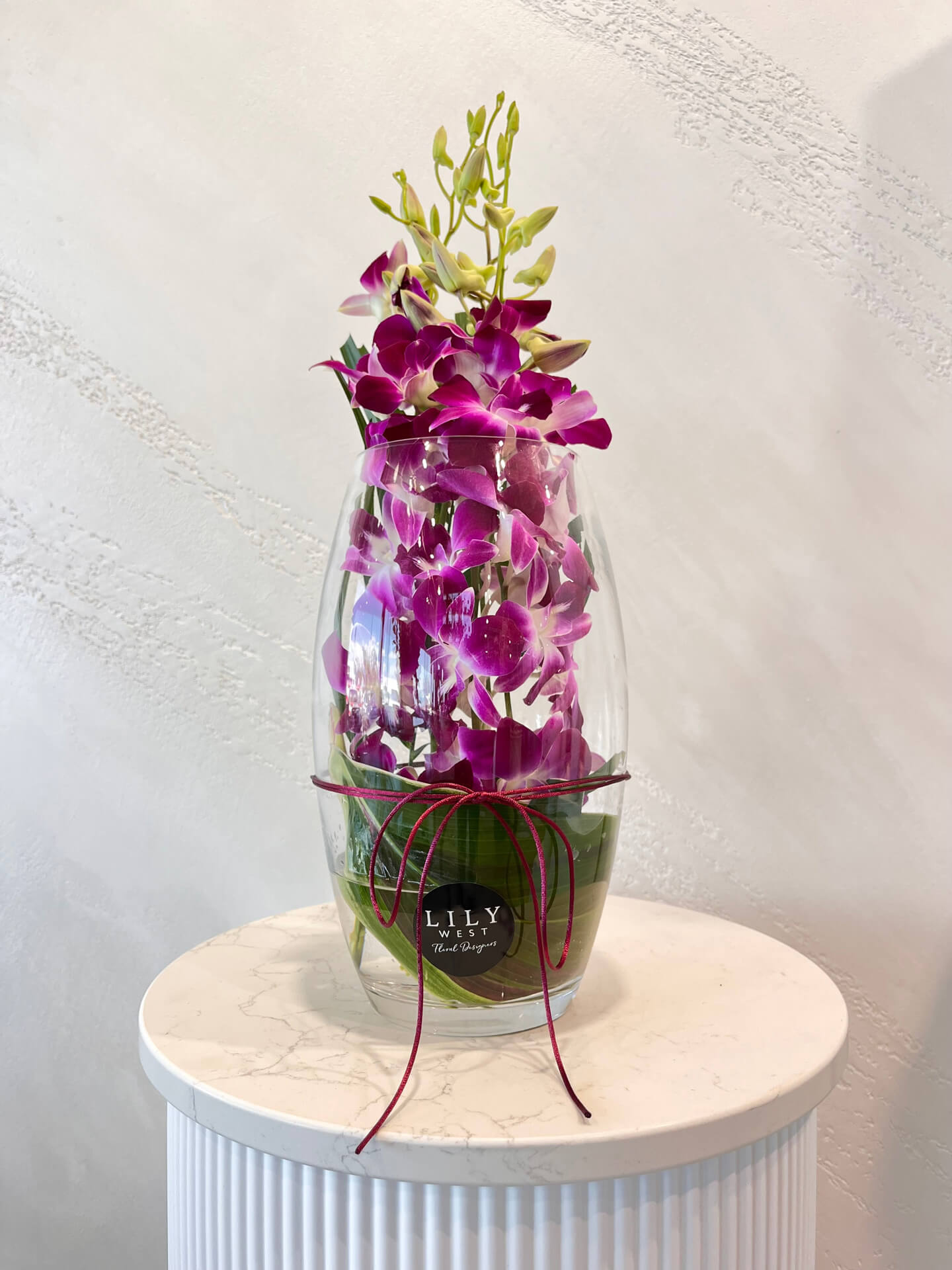 Purple Singapore orchid stems arranged with tropical foliage in a glass vase.