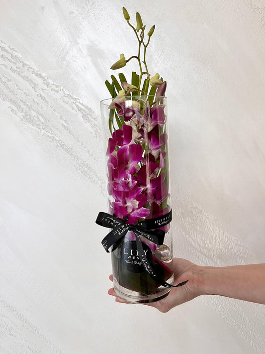Purple Singapore orchid stems arranged with tropical foliage in a glass vase.