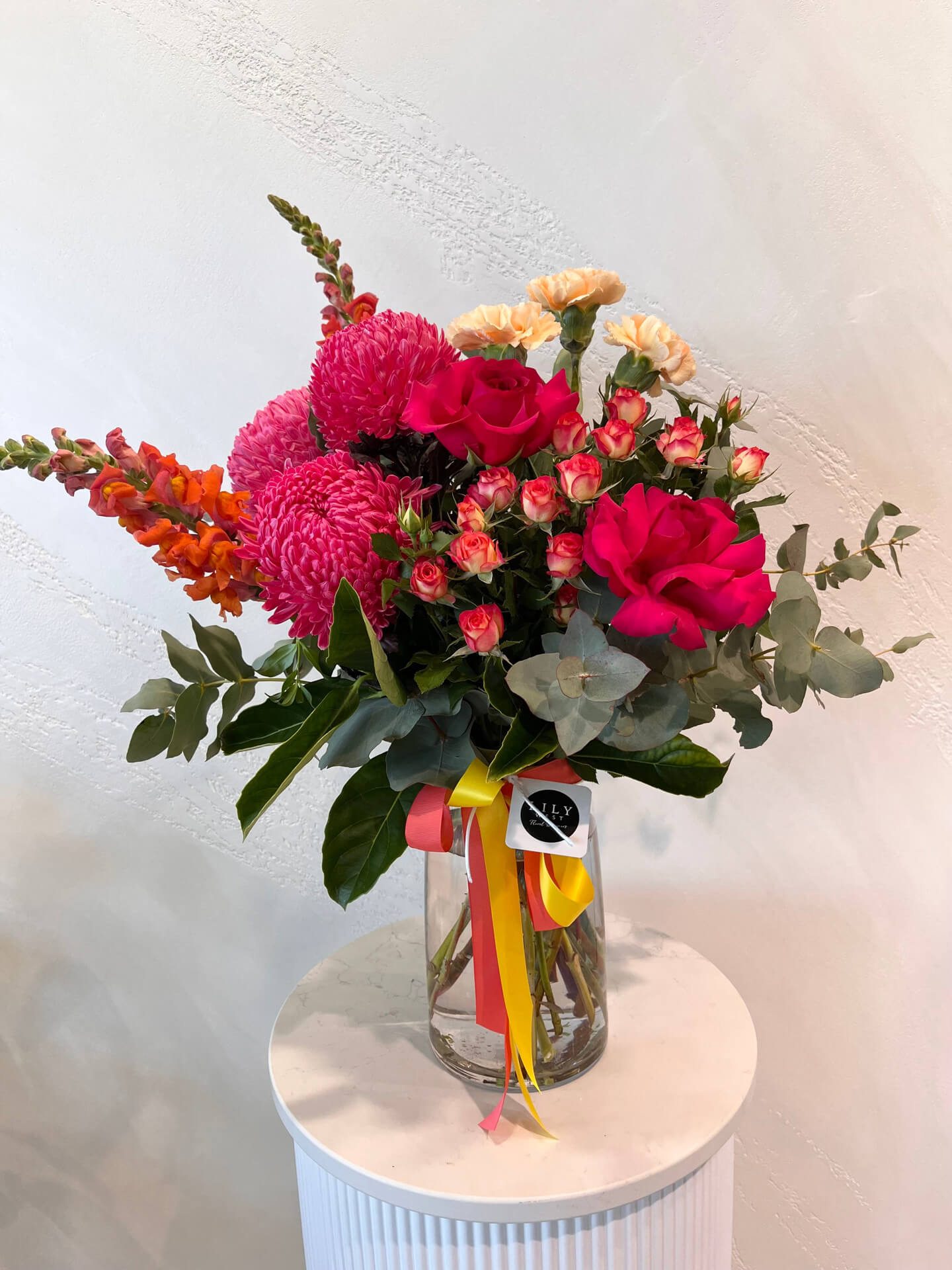 A large bright arrangement of lush foliage, mums, carnations, snapdragons and roses made in a glass vase.