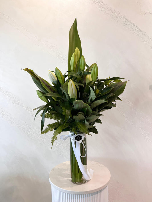 A white oriental lily arrangement with green lush foliage in a tall clear glass vase. 