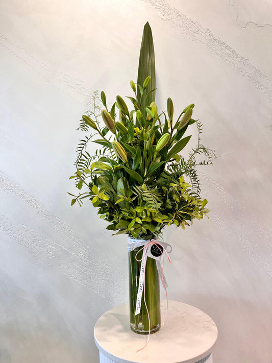A tall arrangement of tiger lilies and lush green foliage in a tall clear glass vase with ribbon.