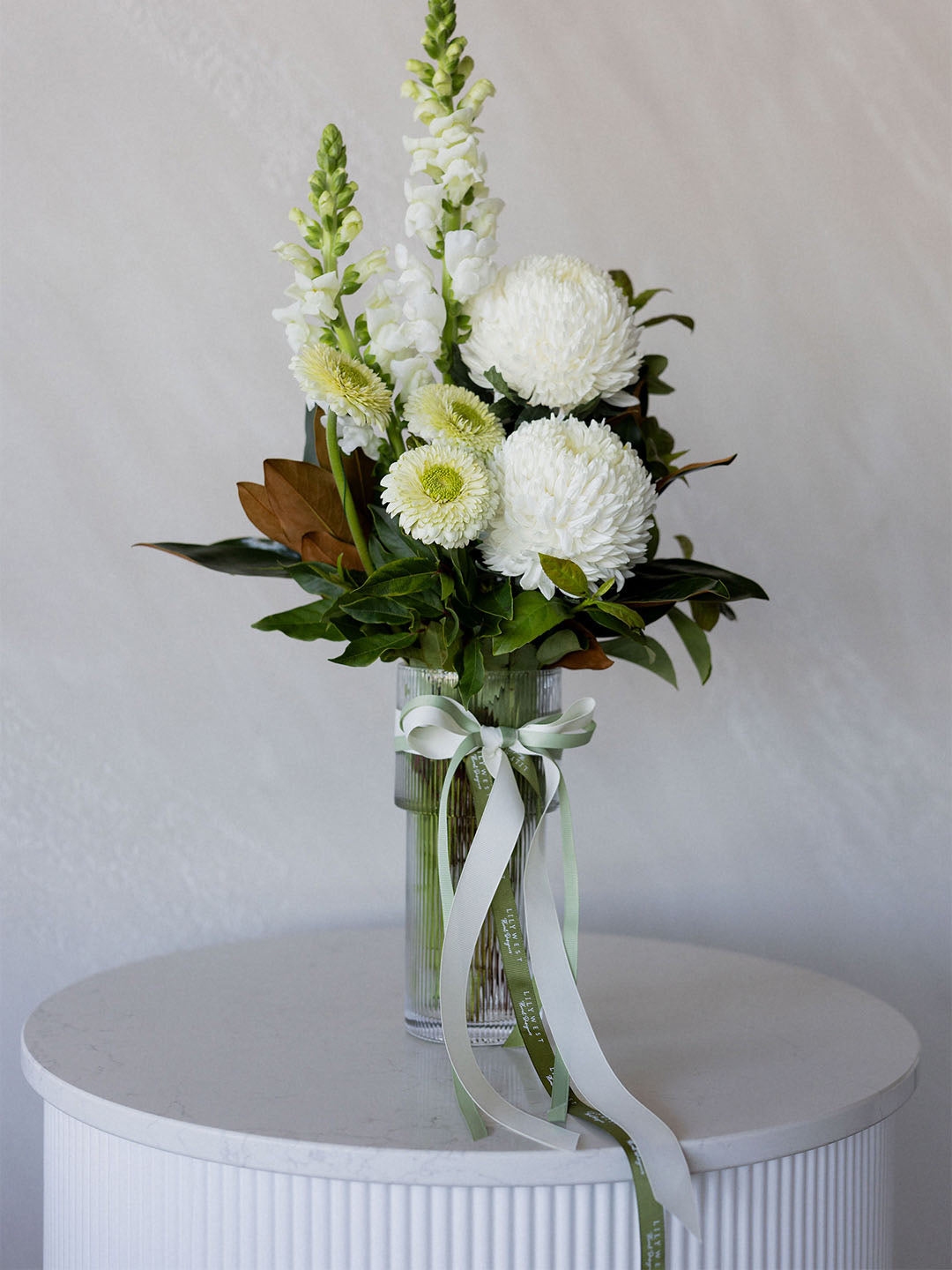 A floral arrangement in a clear glass vase with a pale green ribbon bow, featuring white flowers, green foliage, and a few tan accents, displayed on a white ribbed pedestal against a light gray wall.