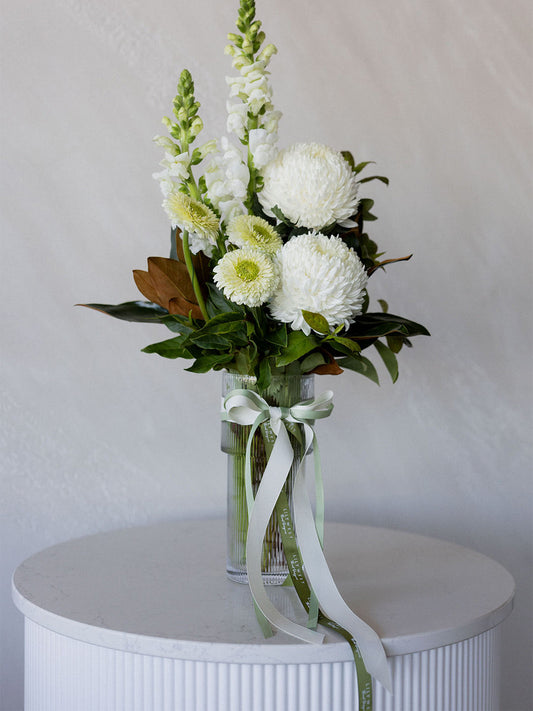 A floral arrangement in a clear glass vase with a pale green ribbon bow, featuring white flowers, green foliage, and a few tan accents, displayed on a white ribbed pedestal against a light gray wall.