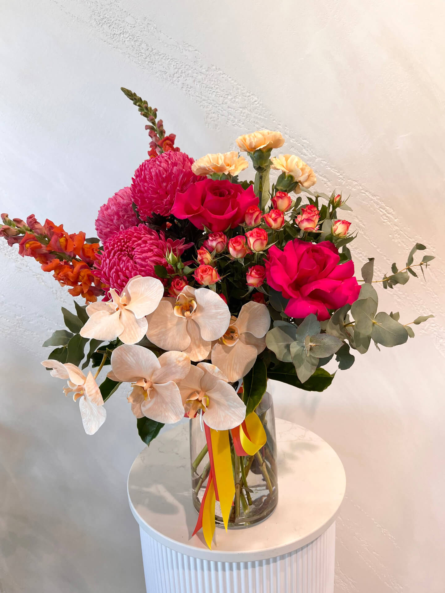 A large bright arrangement of lush foliage, mums, carnations, snapdragons and roses made in a glass vase, upgraded with a phalaenopsis stem. 