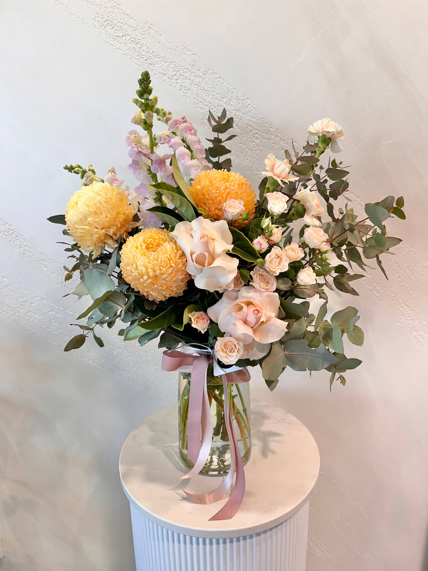 A large pastel arrangement of lush foliage, mums, carnations, snapdragons and roses made in a glass vase.