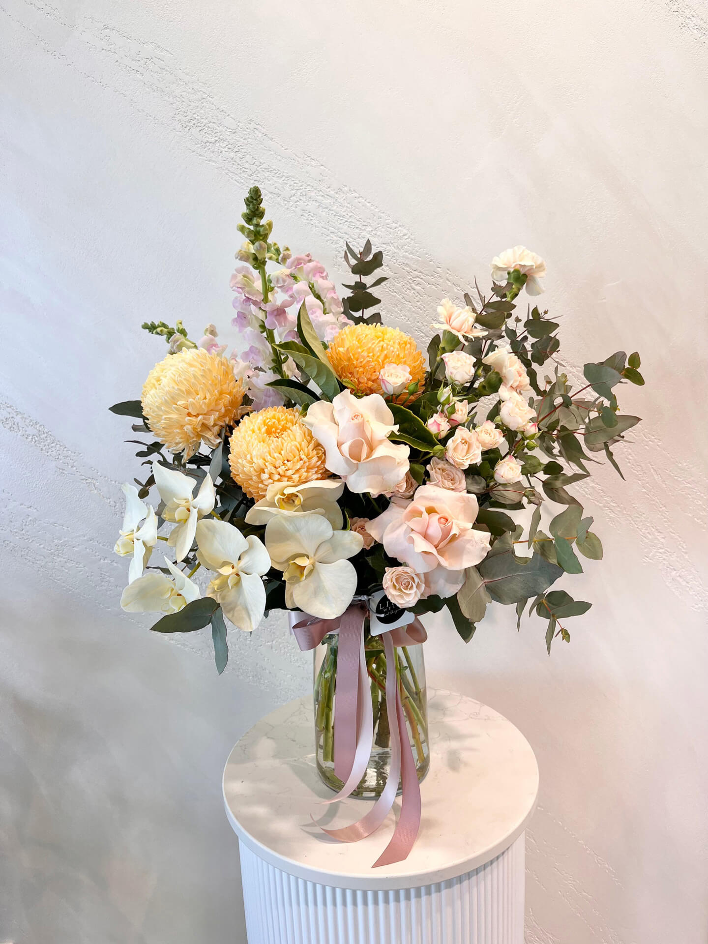 A large pastel arrangement of lush foliage, mums, carnations, snapdragons and roses made in a glass vase.