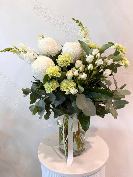 A large white and green arrangement of lush foliage, mums, carnations, snapdragons and roses made in a glass vase.