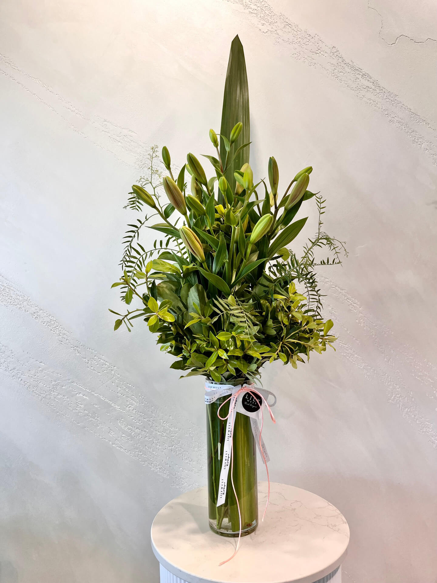 A tall arrangement of tiger lilies and lush green foliage in a tall clear glass vase with ribbon.